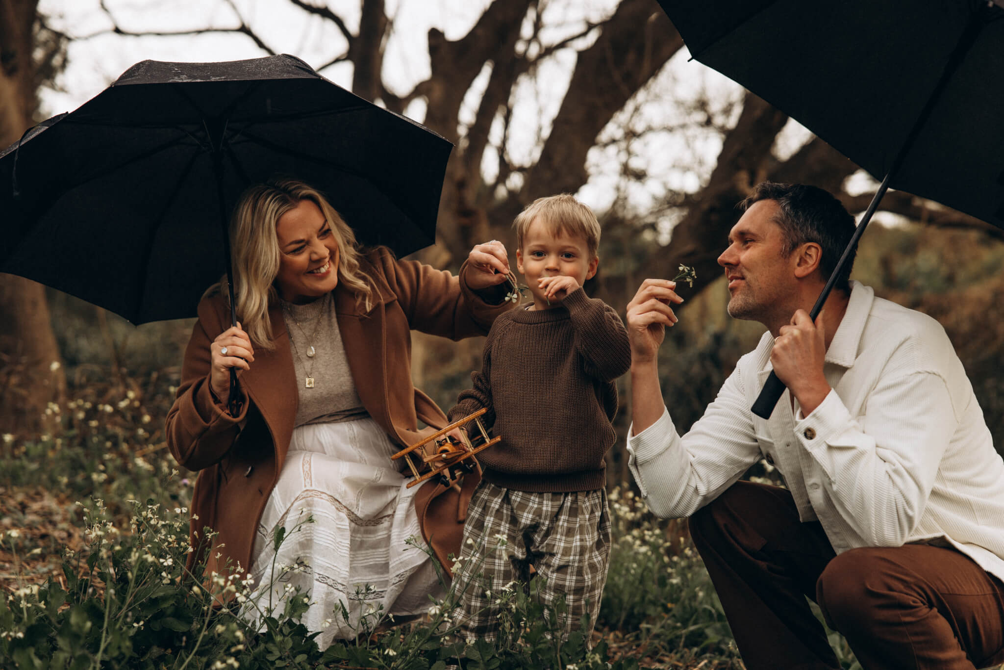 Family photography session in Gympie on a rainy day, parents holding umbrellas and smiling with their young son in the wildflowers.