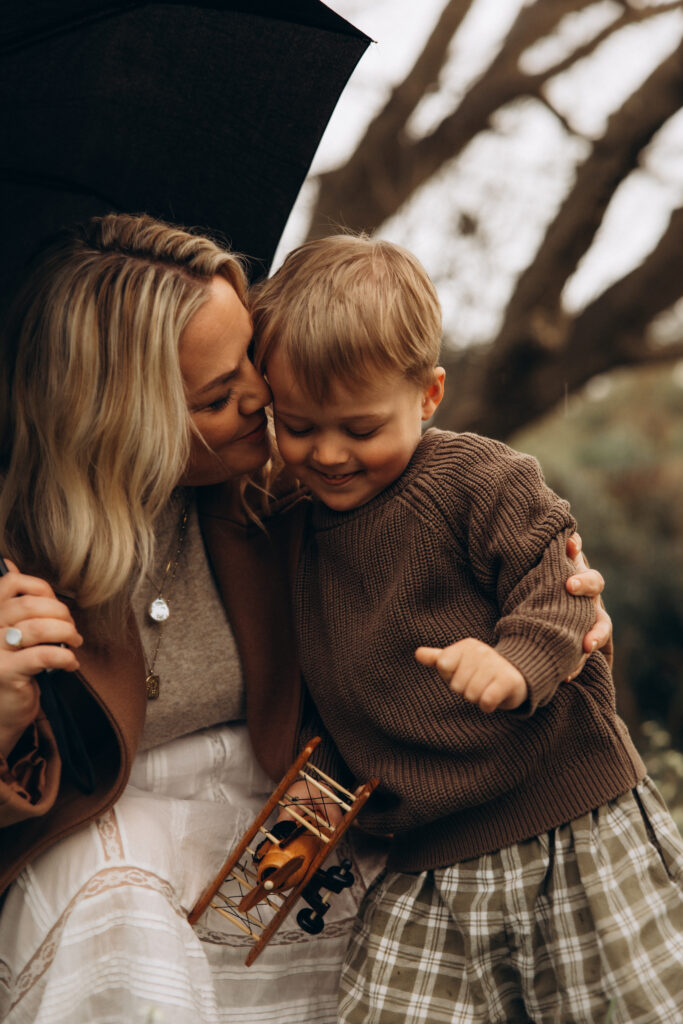 A mother kisses her young son under an umbrella during a rainy family photography in Gympie session, with the boy holding a wooden toy plane.