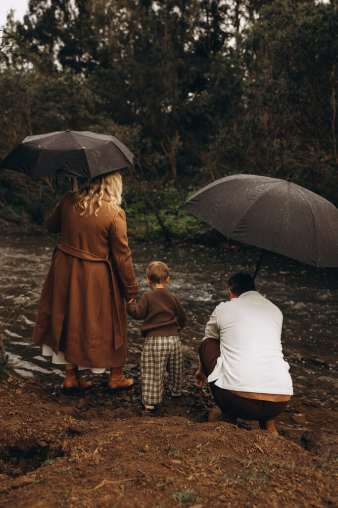 A family stands by the river in Gympie under umbrellas during a rainy photography session, holding hands with their young son. Family photographer in Gympie.