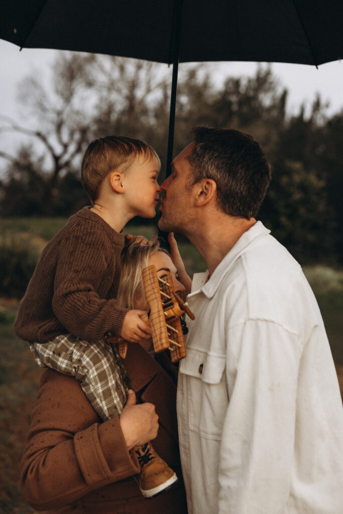 Gympie family photographer. A little boy kisses his father under an umbrella while his mother holds him close during a rainy family photography session in Gympie.