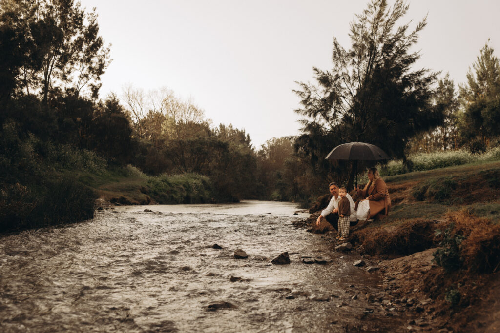 Gympie family photographer. Family sits by the river under an umbrella during a rainy day photography session in Gympie.