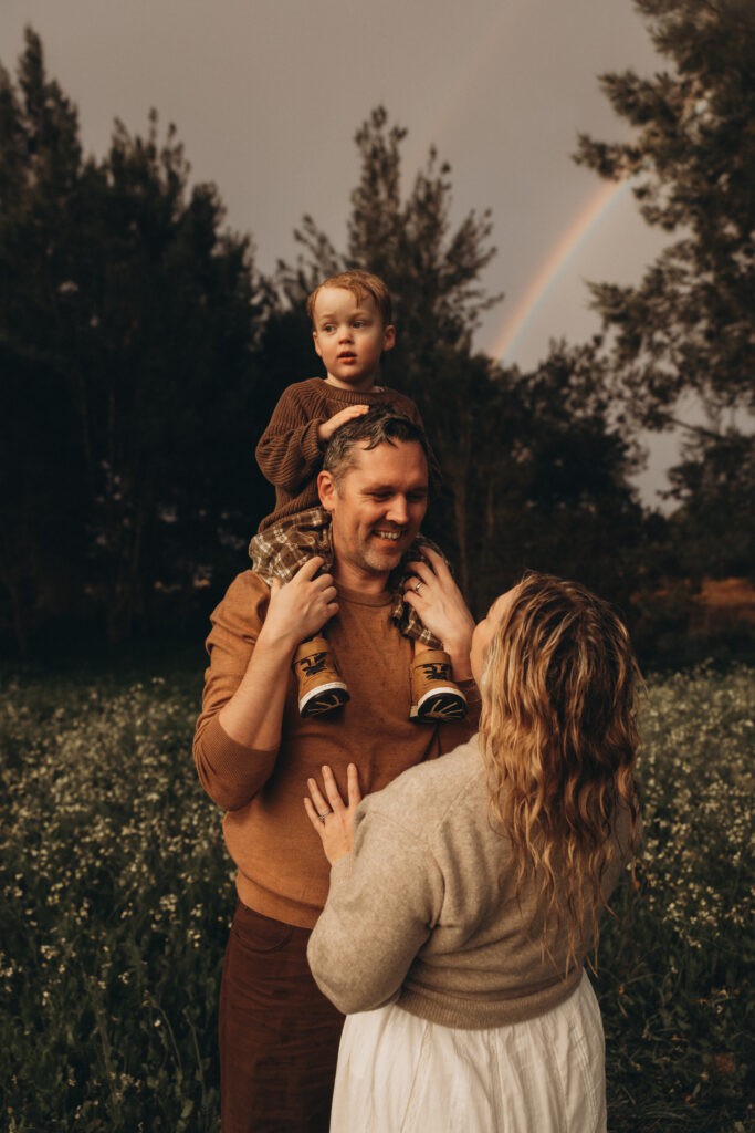 Father carries son on his shoulders while mother looks up at them, with a rainbow arching in the background during a family photoshoot in Gympie.