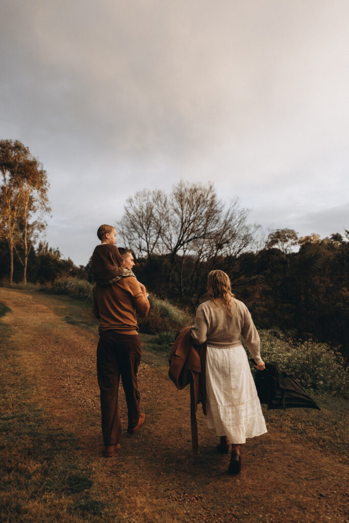 A family walks along a country track in Gympie after the rain, with a child on the father’s shoulders and the mother carrying an umbrella.