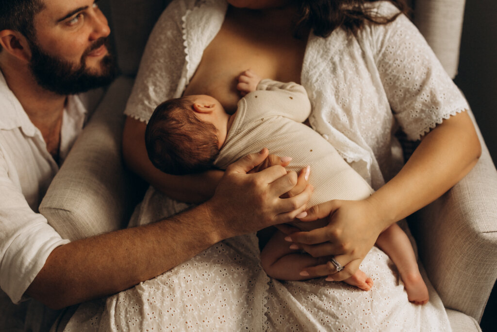 Mother breastfeeding newborn while father holds baby’s leg and looks on lovingly – lifestyle newborn photography Gympie.