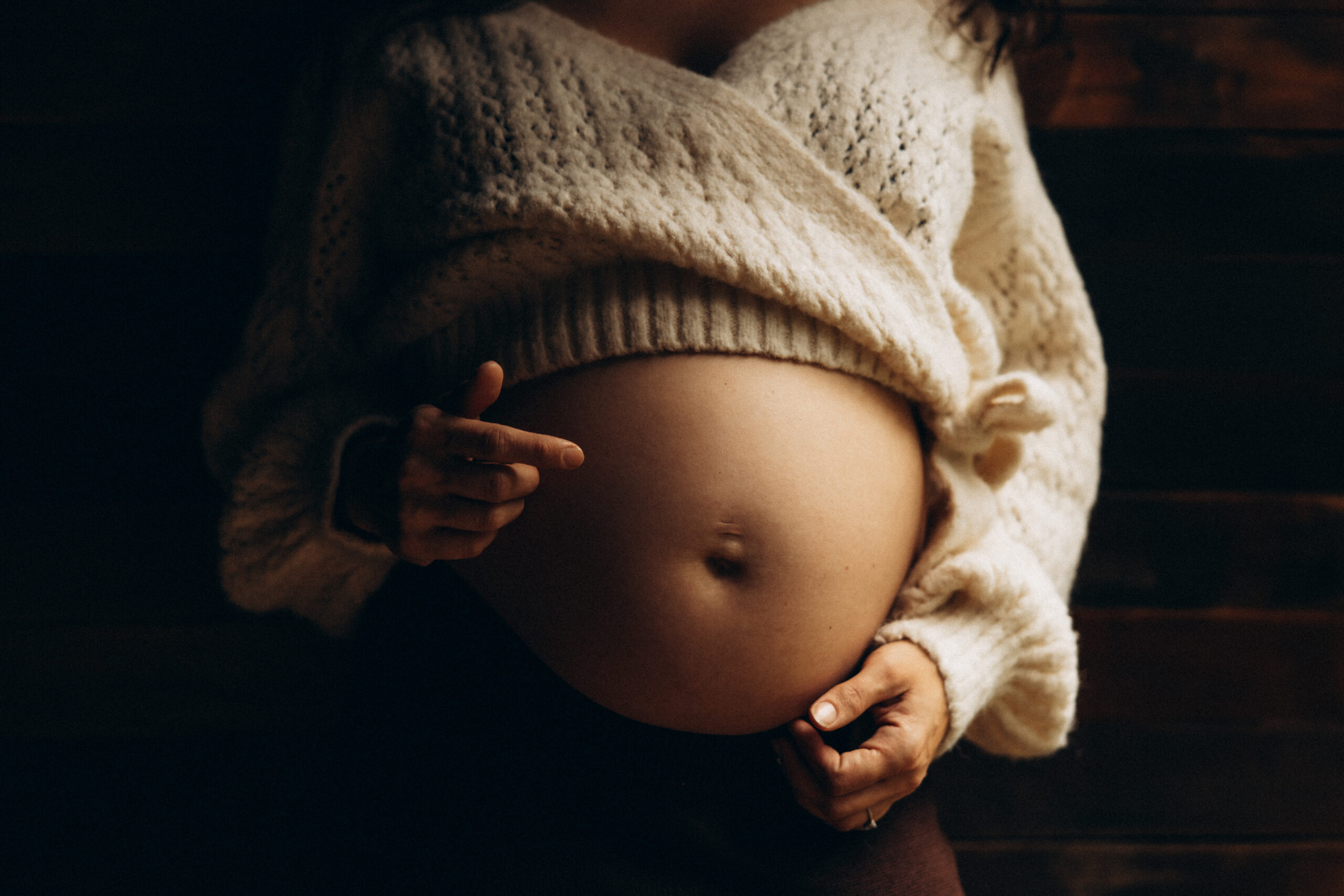 A Gympie maternity photograph of a mother standing by a wooden wall near a window, her pregnant belly softly illuminated by natural light, capturing a soulful moment of motherhood.