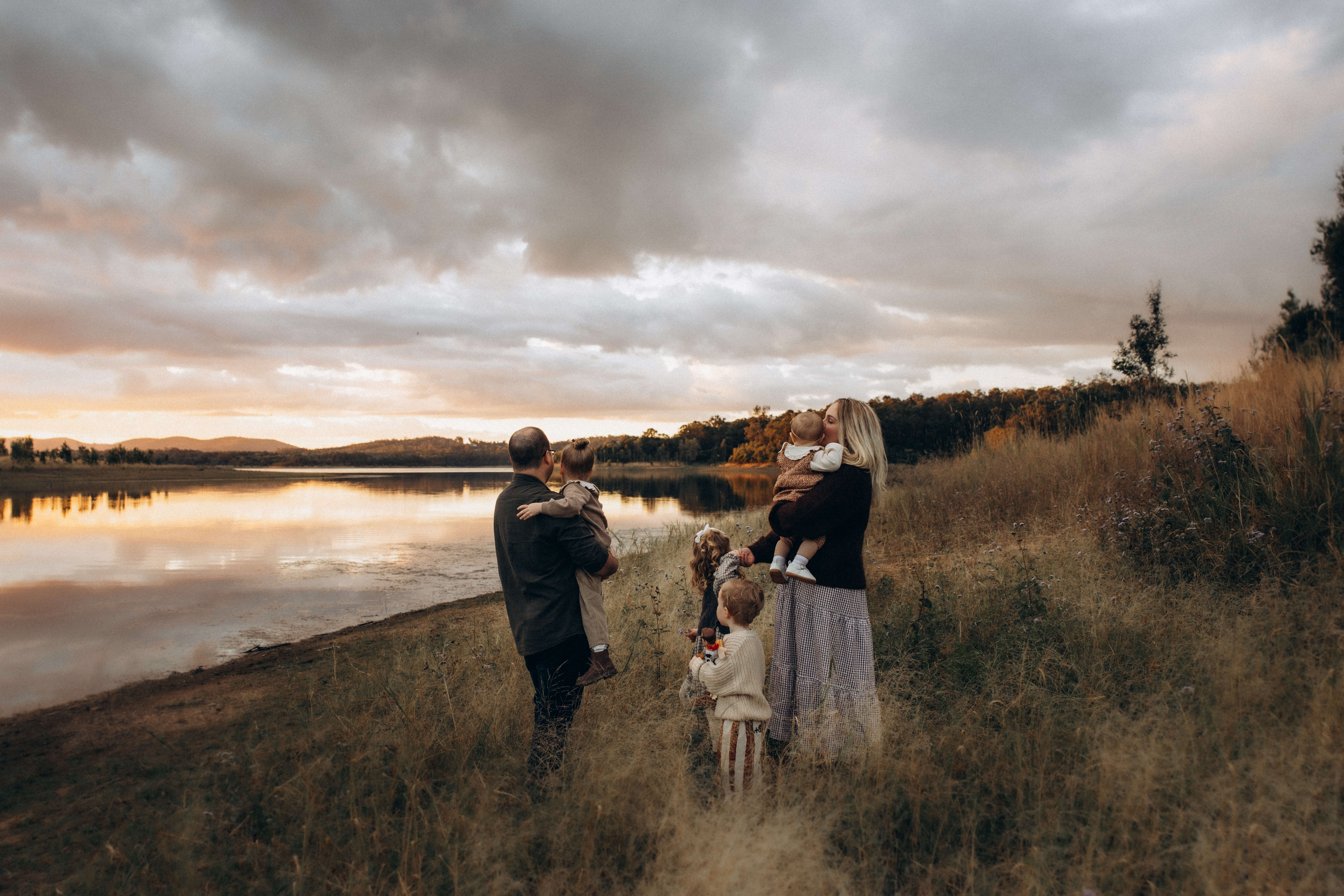 Gympie family photographer. Family of five standing together at sunset by the water in Gympie, captured in warm earthy tones by Rue McConnell Photography.